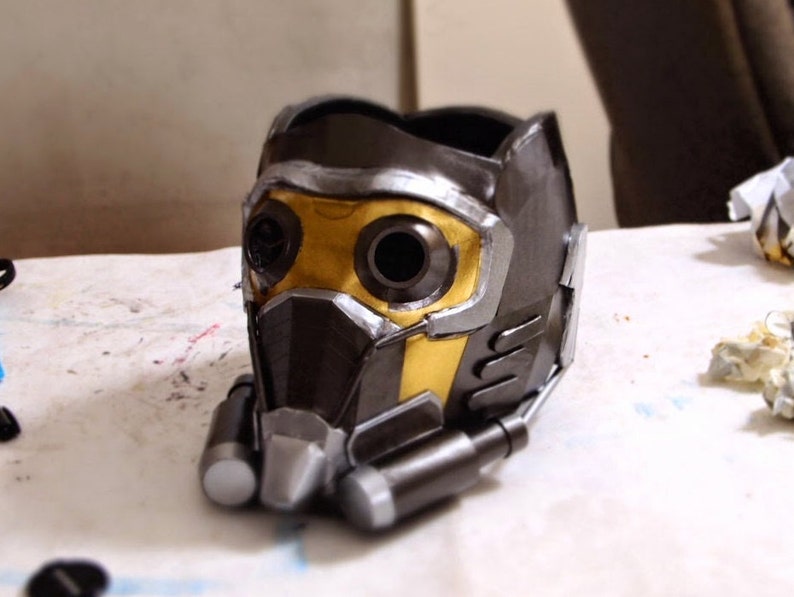 May include: A black and gold Star-Lord helmet with a detailed design. The helmet has two large eye holes and a textured surface.