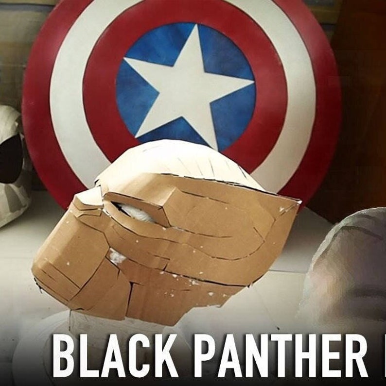 May include: A brown cardboard Black Panther mask is being made. The mask is sitting on a white surface in front of a red, white, and blue Captain America shield.