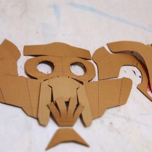 May include: A set of brown cardboard pieces cut out in the shape of a mask. The mask has two eye holes and a detailed design around the nose and mouth.
