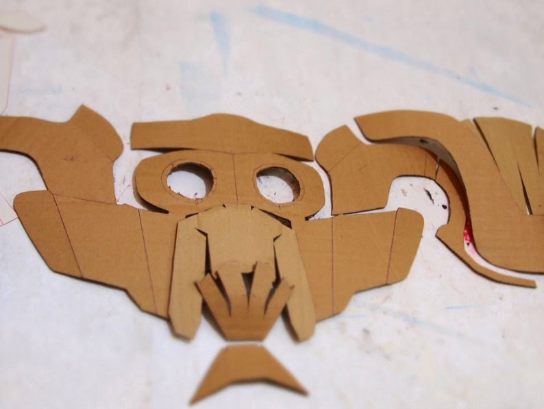 May include: A set of brown cardboard pieces cut out in the shape of a mask. The mask has two eye holes and a detailed design around the nose and mouth.