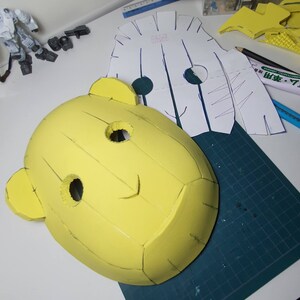 Peut inclure: Un masque en mousse jaune avec un visage souriant, deux grands yeux et deux petites oreilles. Le masque est sur un tapis de coupe vert avec un motif en papier blanc &agrave; c&ocirc;t&eacute;.