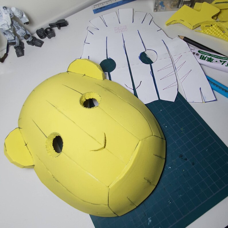 Peut inclure: Un masque en mousse jaune avec un visage souriant, deux grands yeux et deux petites oreilles. Le masque est sur un tapis de coupe vert avec un motif en papier blanc &agrave; c&ocirc;t&eacute;.
