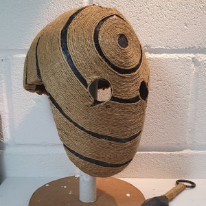 May include: A brown and black mask made of twine with a circular pattern. The mask has two eye holes and a hole on the top.