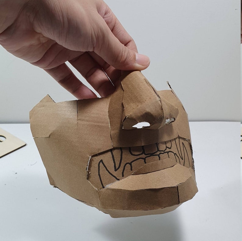 May include: A brown cardboard mask with a detailed design of a face, including a nose, mouth, and teeth. The mask is partially folded, showing the inside.