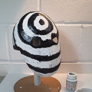 May include: A white and black striped mask with a large black circle in the center. The mask has two eye holes and is made of paper mache.