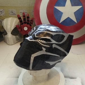 May include: A black and silver Black Panther mask made of paper mache. The mask is on a white mannequin head and is sitting on a white surface.