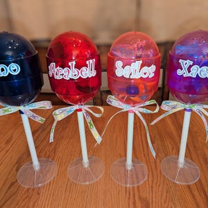 May include: Four clear plastic lollipop holders with white sticks and clear bases. Each holder has a different colored top with a name written on it: Gino, Arabella, Sailor, and Xzela.