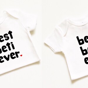 May include: Two white baby onesies with black text. One onesie says "best beti ever" with a small red heart. The other onesie says "best beta ever" with a small red heart.