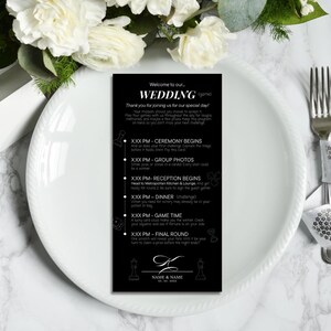 Pode incluir: Um cartão de jogo de casamento em preto e branco com um cronograma de eventos. O cartão apresenta a palavra "WEDDING" no topo, seguida pelos horários e descrições dos eventos. O cartão é colocado em um prato branco com flores no fundo.