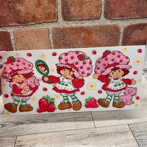 May include: Three Strawberry Shortcake stickers on a white background. Each sticker features Strawberry Shortcake wearing a pink and white dress with red polka dots, a white hat with red strawberries, and green and white striped socks. The stickers are surrounded by small white flowers and red strawberries.