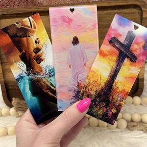 May include: Three rectangular religious art pieces with heart-shaped cutouts at the top. The images depict a helping hand, a figure walking on water, and a cross in a field at sunset. The art pieces are held by a person with pink nail polish.