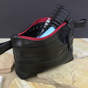 May include: A black zippered pouch made from recycled bicycle inner tubes with a red zipper and a black strap. The pouch is open and contains a hairbrush, a pen, and a blue item.