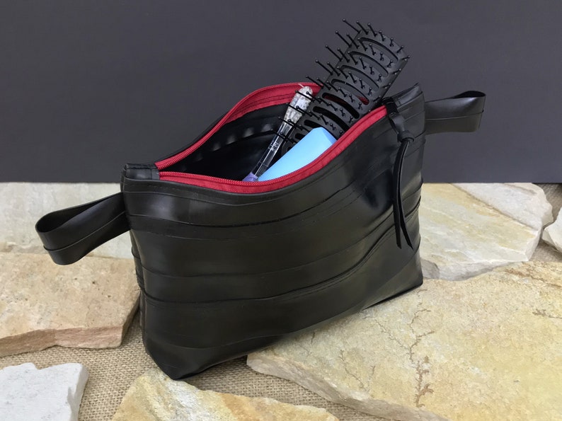 May include: A black zippered pouch made from recycled bicycle inner tubes with a red zipper and a black strap. The pouch is open and contains a hairbrush, a pen, and a blue item.
