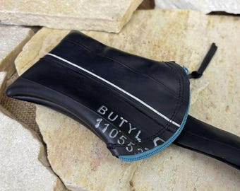 May include: A black zippered pouch crafted from recycled rubber inner tube material. The exterior features a white stripe and the printed text BUTYL 11055. A light blue zipper track runs along the curved edge, complemented by a black fabric zipper pull.