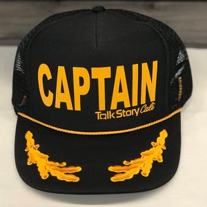 May include: Black trucker hat with the word "CAPTAIN" in gold block letters. The hat features a gold rope trim and two gold embroidered leaf designs on the front. The text "Talk Story Cali" is below the word "CAPTAIN".