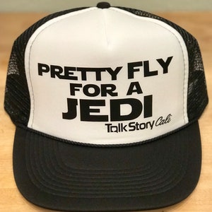 May include: Black and white trucker hat with the text "PRETTY FLY FOR A JEDI" in bold, black letters. The hat has a black brim and mesh back, with "TalkStory Cali" printed below the main text.