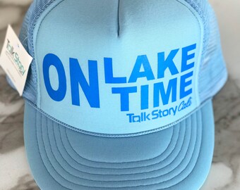 May include: A light blue trucker hat with a mesh back. The front of the hat has the words "ON LAKE TIME" in blue lettering with the words "Talk Story Cafe" in smaller lettering below.