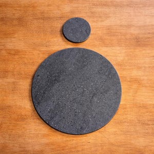 May include: Two dark grey, round stone serving pieces on a wooden surface. The larger piece is a serving platter, and the smaller piece is a coaster. The stone has a speckled texture.