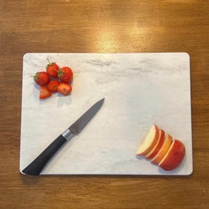 Op de afbeelding: Een rechthoekige marmeren snijplank met aardbeien en gesneden appel. Een roestvrijstalen mes met een zwart handvat ligt op de plank. De plank heeft een wit en grijs patroon.