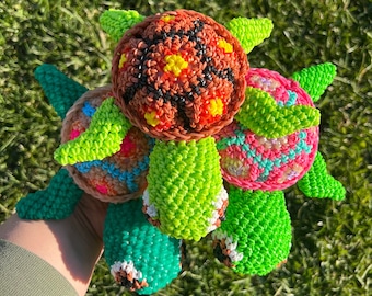 May include: A collection of colorful, handmade rubber band turtles. Each turtle features a round body with a unique pattern of colors, including brown, orange, yellow, pink, and green. The turtles have green flippers and are set against a backdrop of green grass.