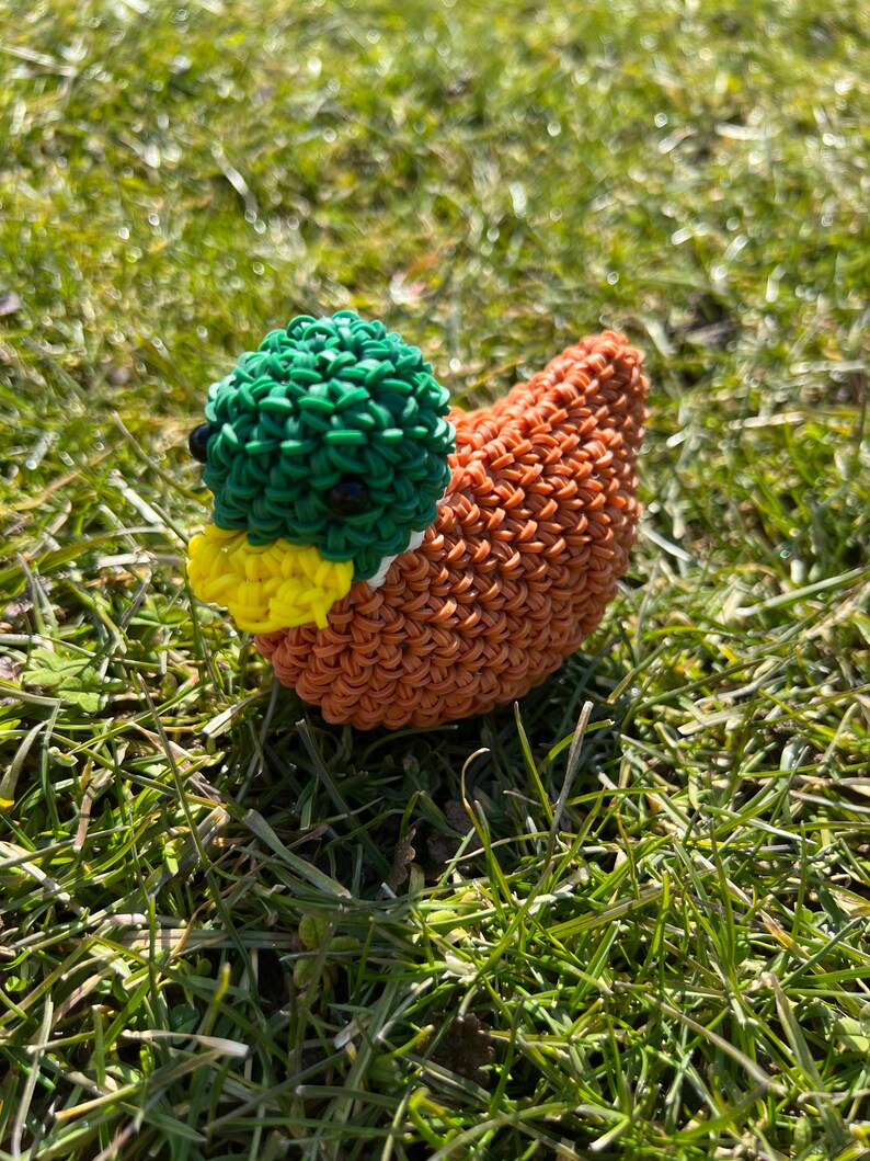 Op de afbeelding: Een groene, gele en oranje rubberen band eend. De eend zit op een bed van groen gras.