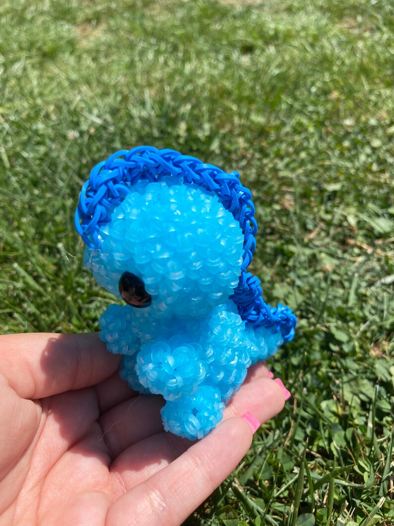 May include: A blue rubber band dinosaur toy with a blue rubber band mane. The dinosaur is made of small rubber bands and has a black eye.