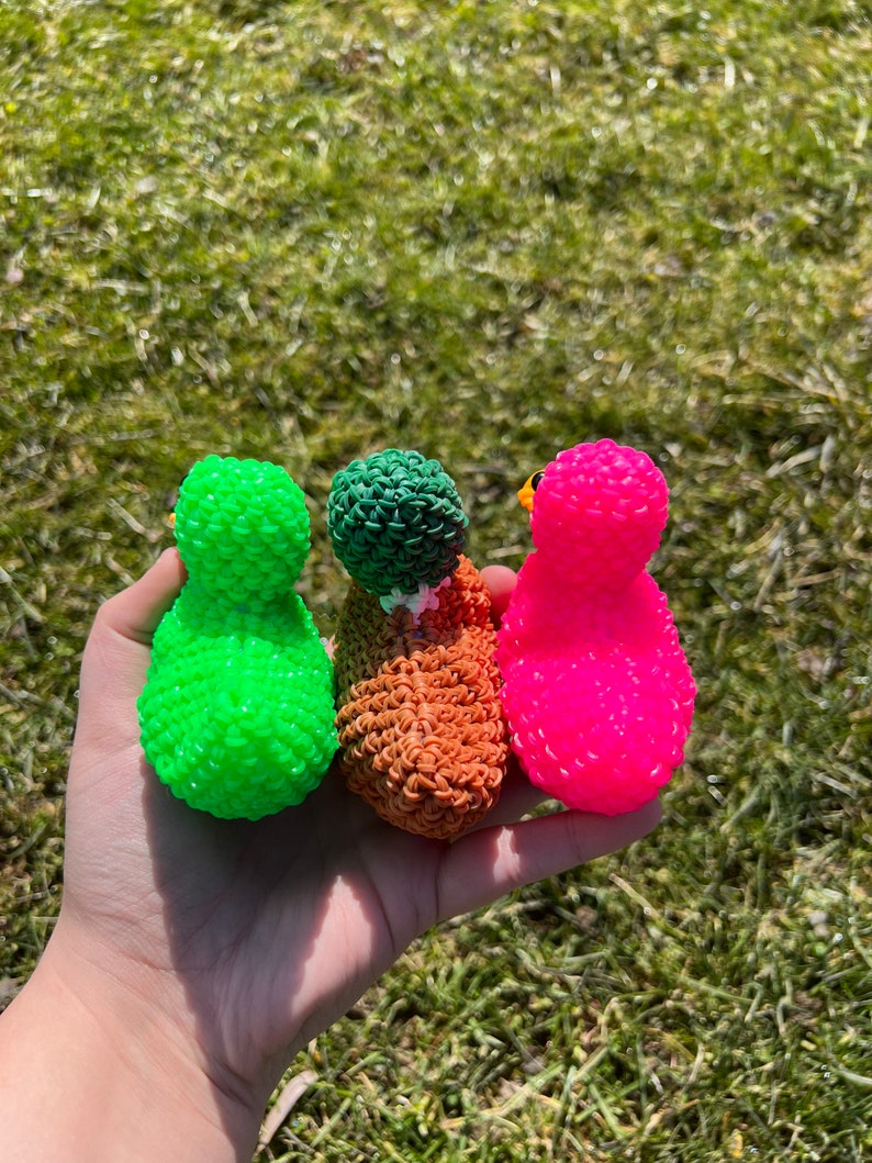 Op de afbeelding: Drie rubberband loom creaties in de vorm van eenden. De eenden zijn groen, oranje en roze.