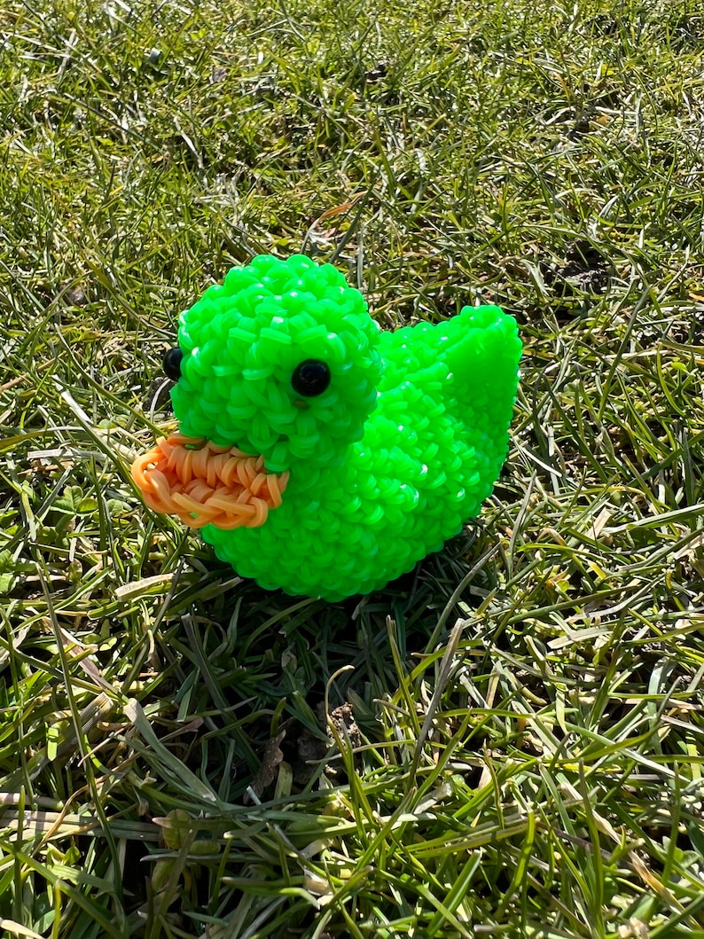 Op de afbeelding: Een groene rubberen eend met zwarte ogen en een oranje snavel. De eend zit op een bed van groen gras.