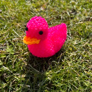 Op de afbeelding: Een roze rubberen eend met een gele snavel en zwarte ogen. De eend zit op een bed van groen gras.