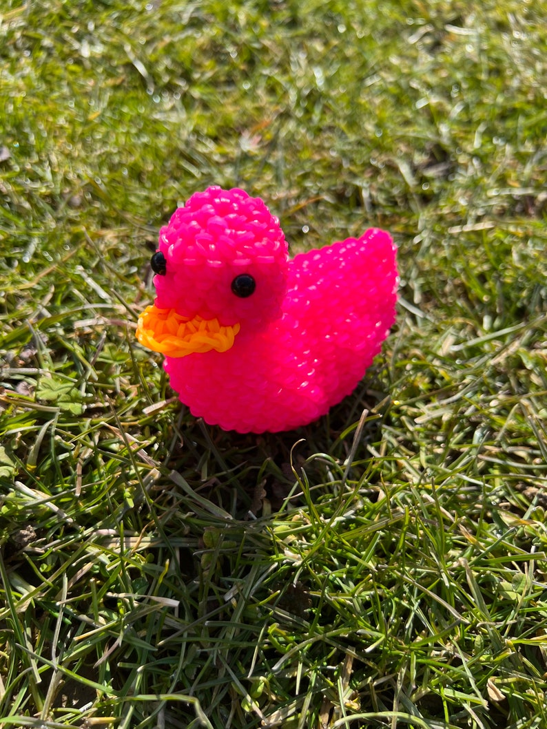 Op de afbeelding: Een roze rubberen eend met een gele snavel en zwarte ogen. De eend zit op een bed van groen gras.