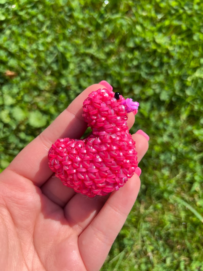 May include: A pink rubber band duck with a purple beak and black eyes. The duck is made of many small rubber bands woven together.