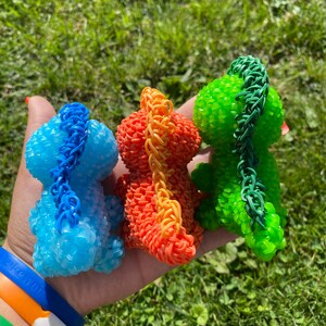 May include: Three small dinosaur figurines made of colorful rubber bands. The dinosaurs are blue, orange, and green.