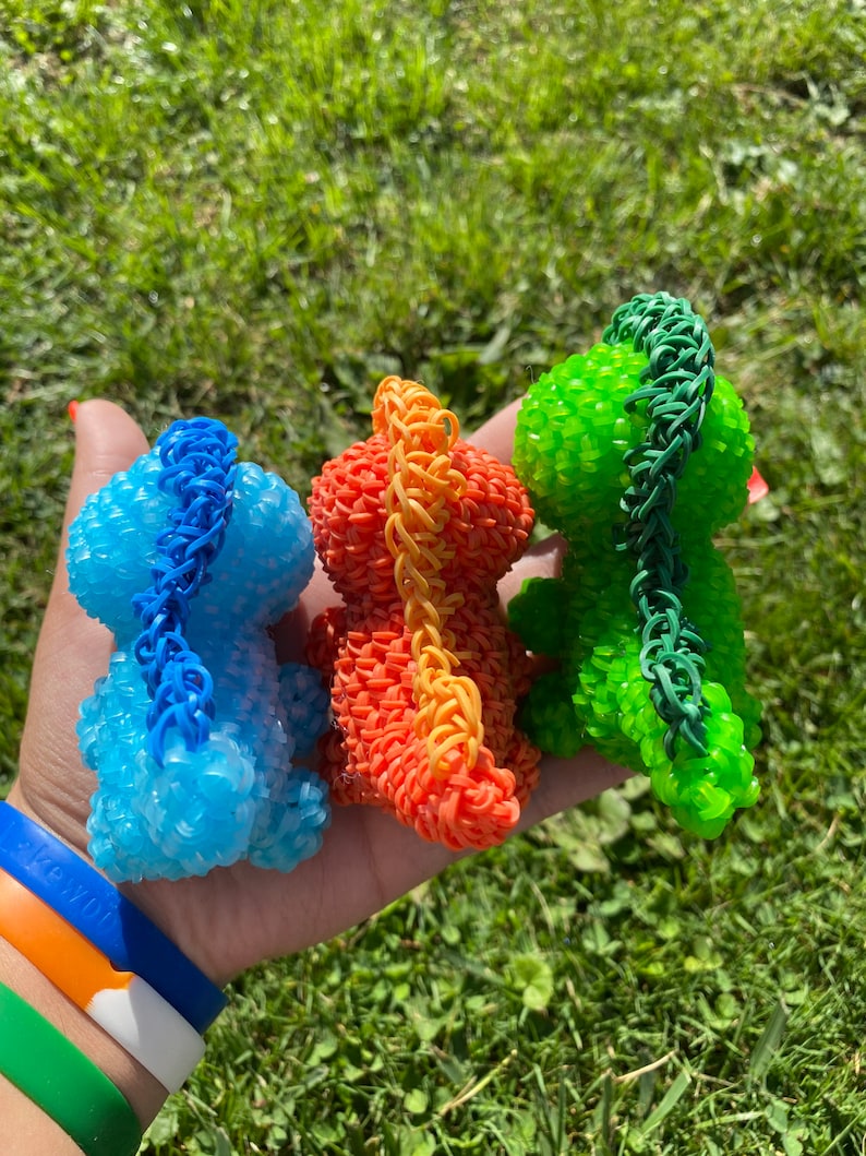 May include: Three small dinosaur figurines made of colorful rubber bands. The dinosaurs are blue, orange, and green.