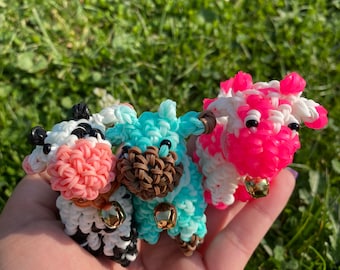 May include: Three small cow figurines made of colorful rubber bands. The cows are black and white, pink and white, and blue and brown. Each cow has a small gold bell hanging from its neck.