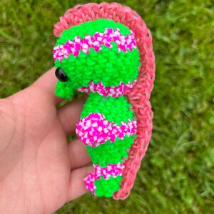 Puede incluir: Un caballito de mar de gomas el&aacute;sticas verde y rosa con un ojo negro. El caballito de mar est&aacute; hecho con gomas el&aacute;sticas de telar.