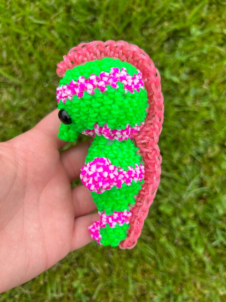 Puede incluir: Un caballito de mar de gomas el&aacute;sticas verde y rosa con un ojo negro. El caballito de mar est&aacute; hecho con gomas el&aacute;sticas de telar.