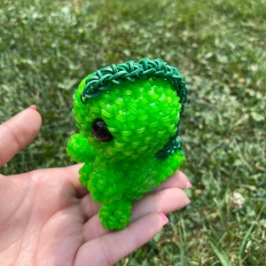 May include: A green rubber band toy in the shape of a small dinosaur with a green rubber band mane. The toy has large black eyes and is being held in a person's hand.