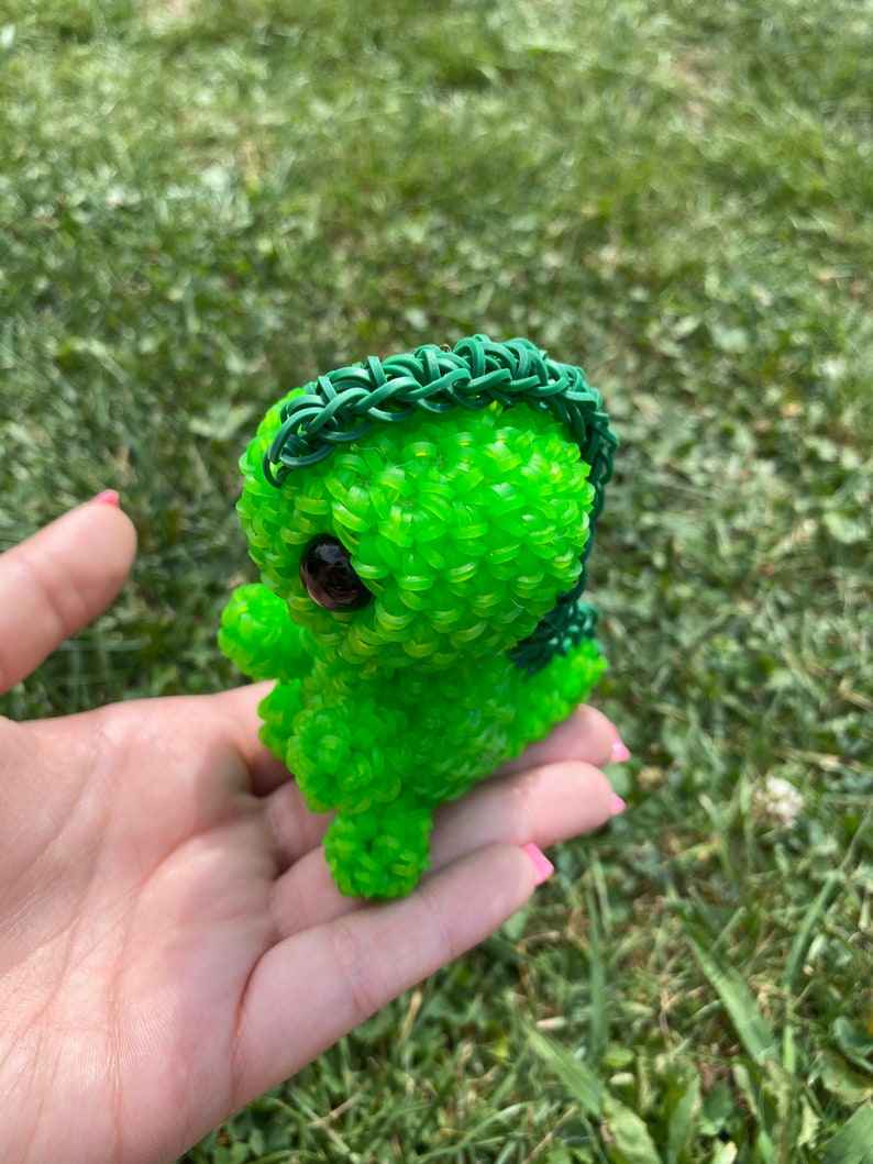 May include: A green rubber band toy in the shape of a small dinosaur with a green rubber band mane. The toy has large black eyes and is being held in a person's hand.