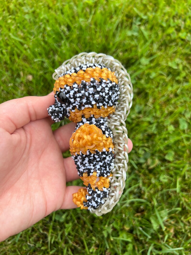 Puede incluir: Una peque&ntilde;a figura de caballito de mar hecha a mano con gomas el&aacute;sticas de colores. El caballito de mar es negro, blanco y naranja con una cola de goma el&aacute;stica blanca y transparente.