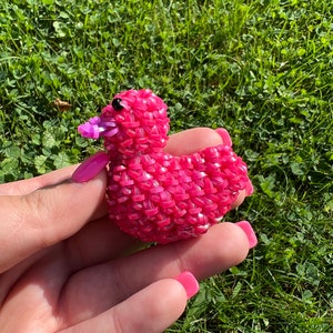 May include: A pink rubber band duck with a purple beak and black eyes. The duck is sitting in a person's hand.