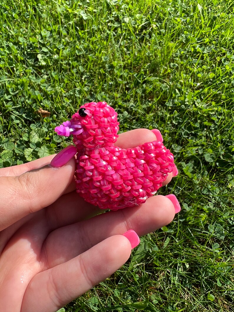May include: A pink rubber band duck with a purple beak and black eyes. The duck is sitting in a person's hand.