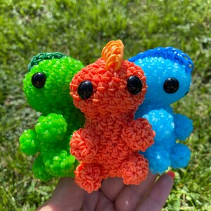 May include: Three small crocheted plushies in the shape of a dinosaur. The plushies are green, orange, and blue.