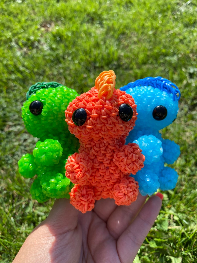 May include: Three small crocheted plushies in the shape of a dinosaur. The plushies are green, orange, and blue.