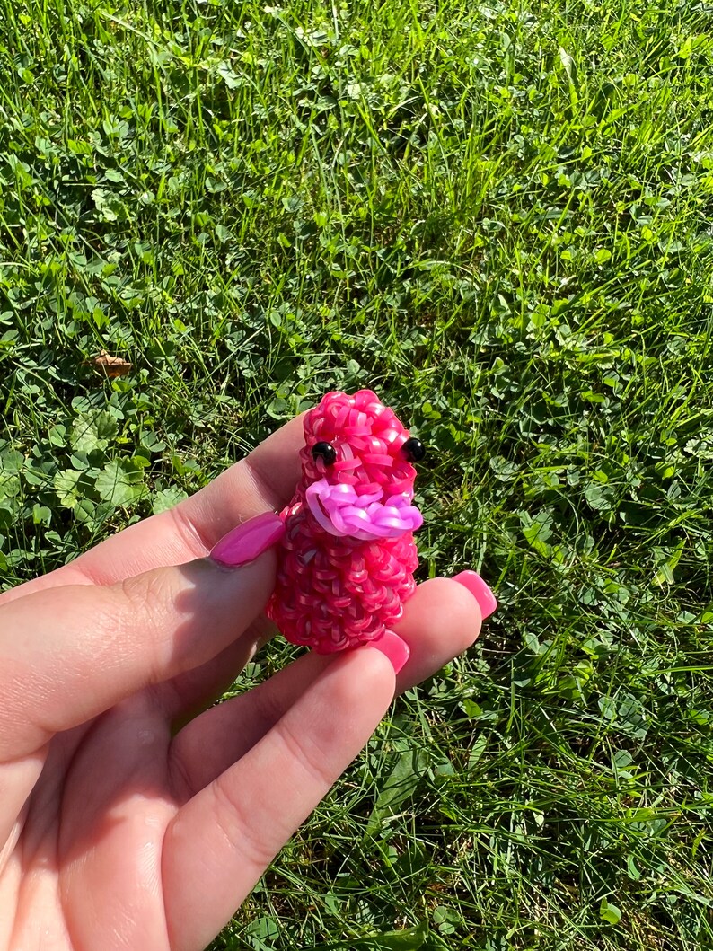 May include: A pink rubber band animal with black eyes and a pink mouth. The animal is made of rubber bands and is about the size of a small apple.