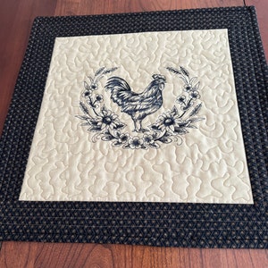May include: A quilted placemat featuring a rooster design. The center has a cream-colored background with a black rooster illustration surrounded by flowers. The border is black with a repeating pattern.