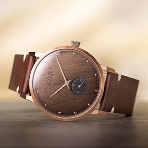 May include: A rose gold wristwatch with a brown wooden face and a small seconds dial. The watch has a brown leather band. The watch face has the text "AVANT NEW YORK".