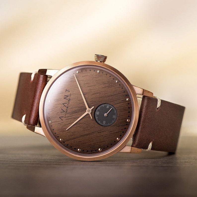 May include: A rose gold wristwatch with a brown wooden face and a small seconds dial. The watch has a brown leather band. The watch face has the text "AVANT NEW YORK".