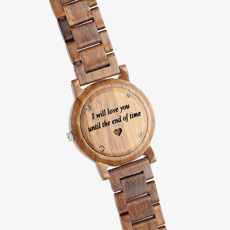 Puede incluir: Un reloj de pulsera de madera con una banda marr&oacute;n y una cara redonda. La cara tiene un coraz&oacute;n recortado y un mensaje grabado que dice "Te amar&eacute; hasta el fin del tiempo".