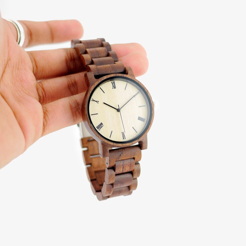 Puede incluir: Un reloj de pulsera de madera marr&oacute;n con una esfera de color crema y marcadores de hora romanos negros. El reloj tiene una correa de madera marr&oacute;n.
