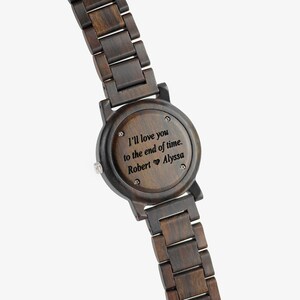 Puede incluir: Un reloj de pulsera de madera marr&oacute;n oscuro con una esfera redonda y un mensaje grabado en negro que dice "Te amar&eacute; hasta el final de los tiempos. Robert &hearts; Alyssa".